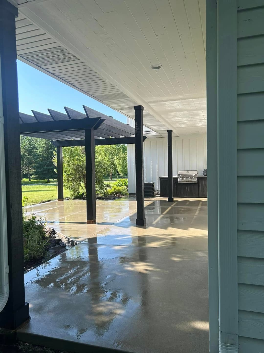Modern patio with black beams, polished concrete floor, and grill area surrounded by greenery.