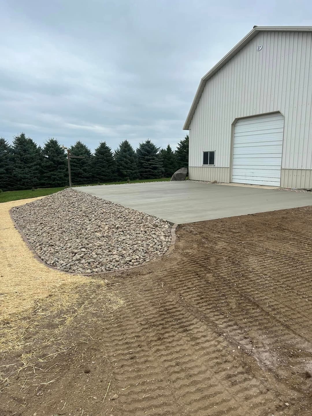 Newly poured concrete pad beside a rural storage building with landscaping stones and grass.