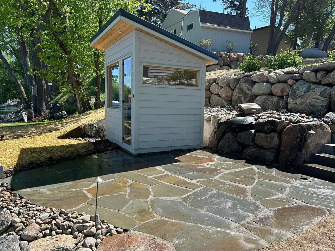 Modern garden shed beside stone patio and lush greenery in a serene outdoor setting.