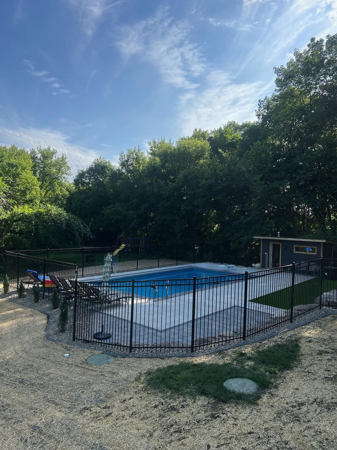 Backyard swimming pool surrounded by a fence and trees, with lounge chairs and landscaping.