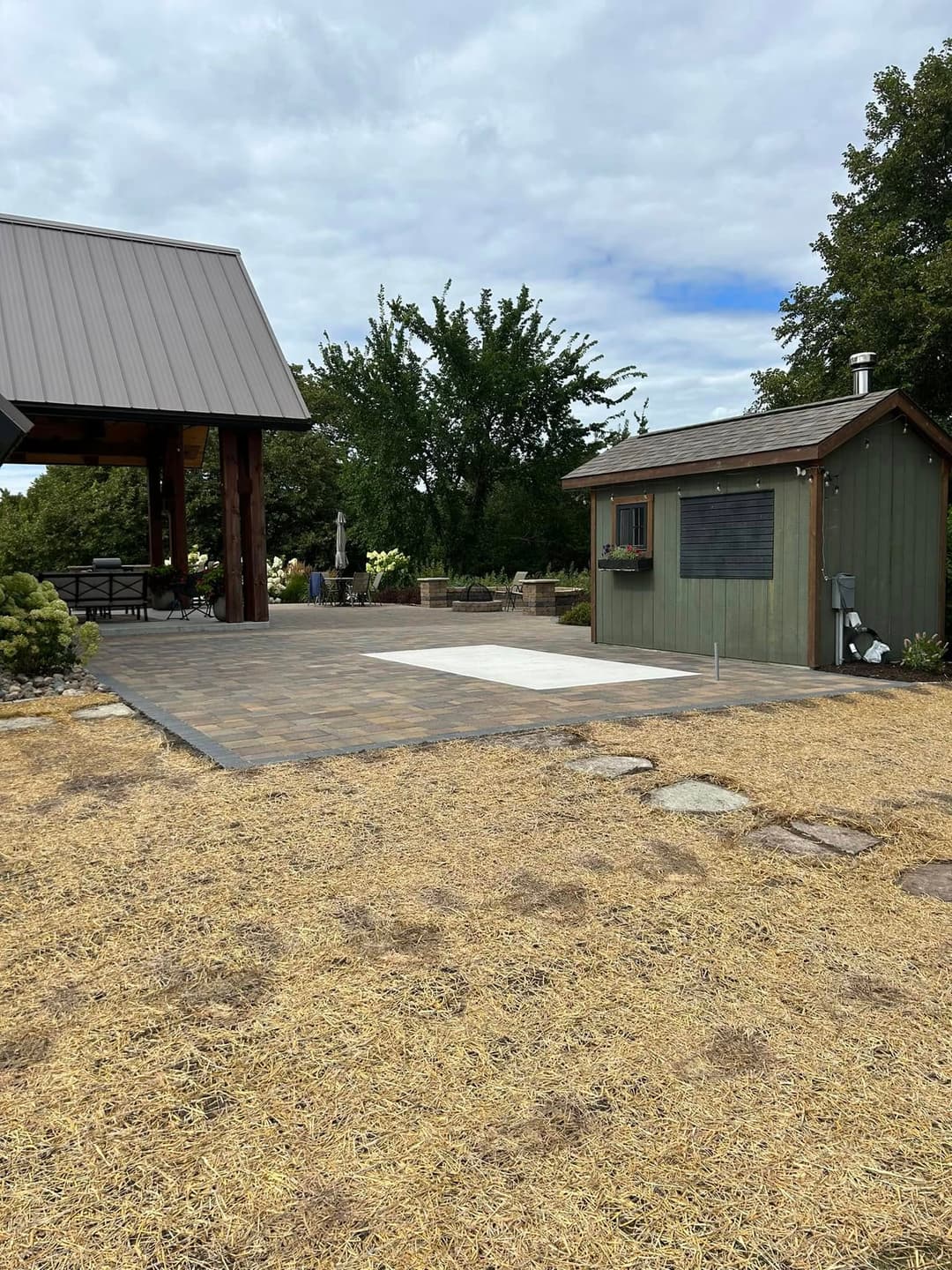 Outdoor patio area with a stone surface and a green shed surrounded by dry grass and trees.