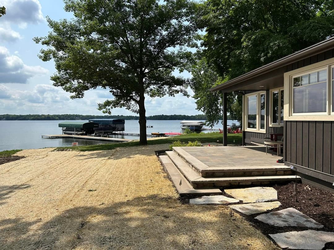 Lakeside view of cottage with patio, trees, and docked boats under a blue sky.
