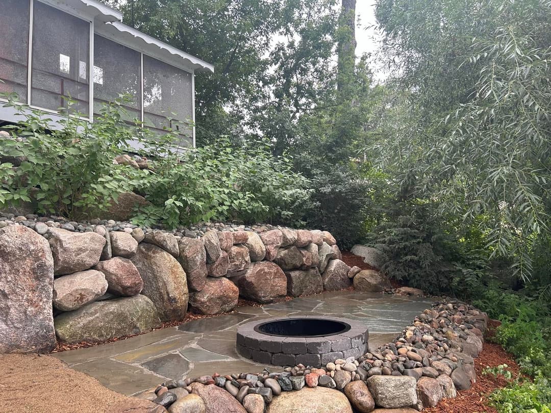 Stone patio with circular fire pit surrounded by lush greenery and boulders.