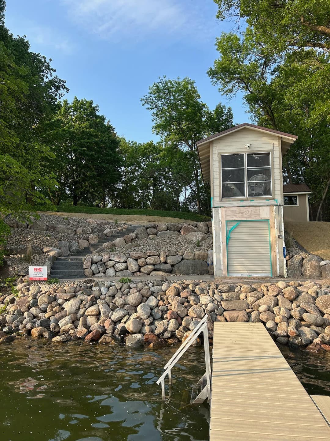 Lakeside view of a stone wall and boat dock with a storage shed among green trees.