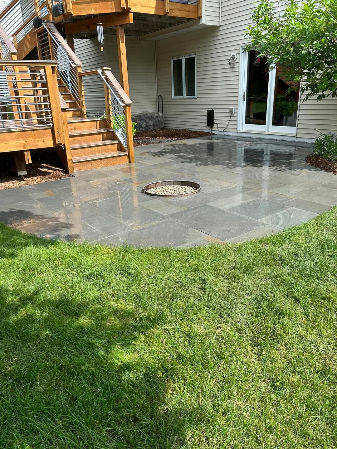 Patio area with slate tiles and a circular stone feature near a wooden deck and grass.