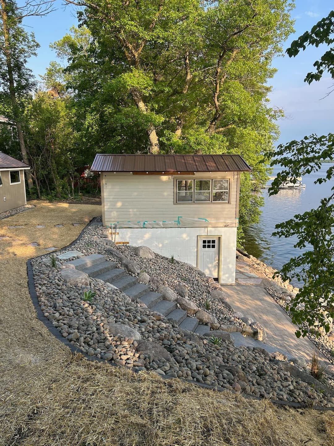Small lakeside cabin with a metal roof, stone pathways, and surrounded by trees.