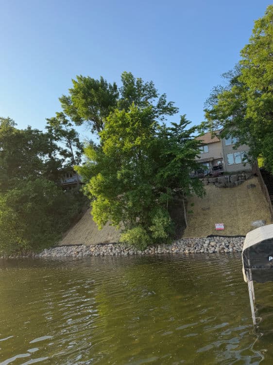 House on a hillside by a lake surrounded by lush trees and rocky shoreline.