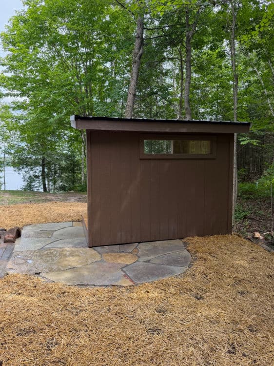 Brown shed nestled in a wooded area with stone pathway and lake view in the background.