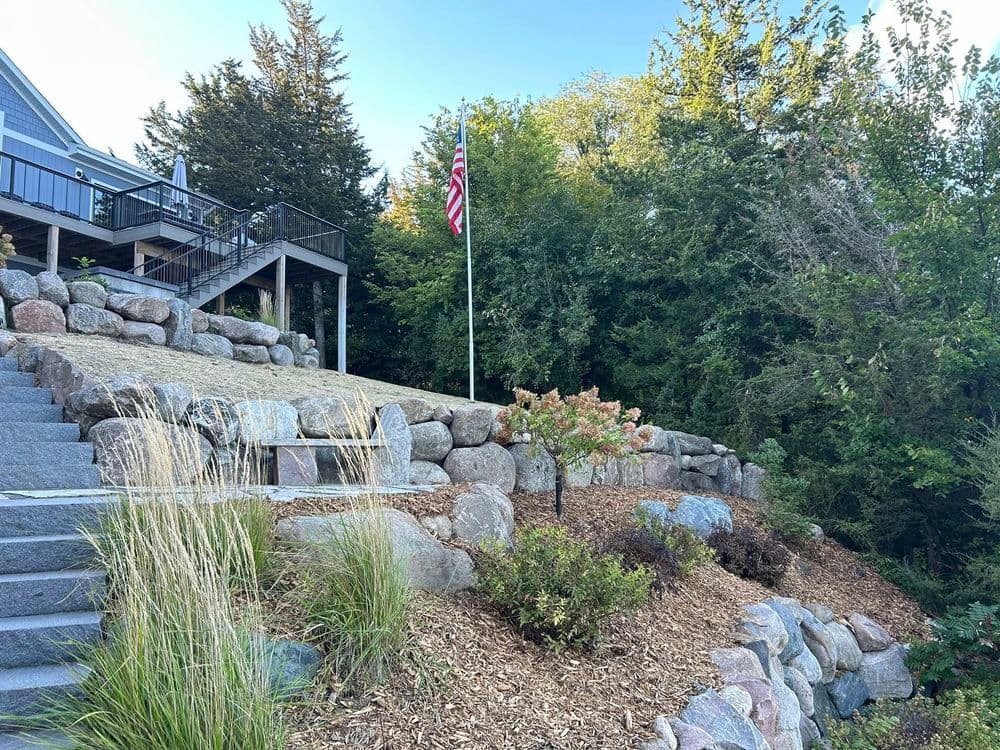 Terraced landscape with stone steps, American flag, and lush greenery near a house.